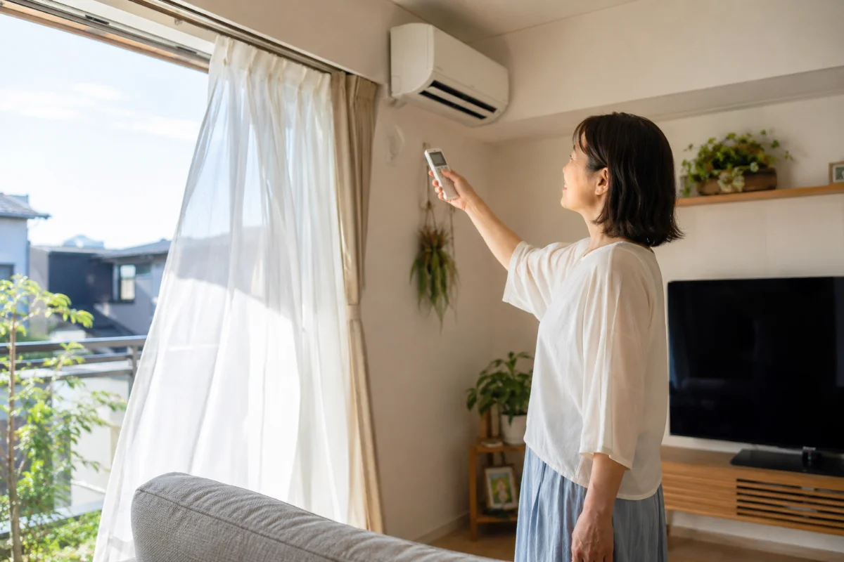 窓を開けて換気しながらエアコンのリモコンを操作し、最低温度の冷房運転を試す女性