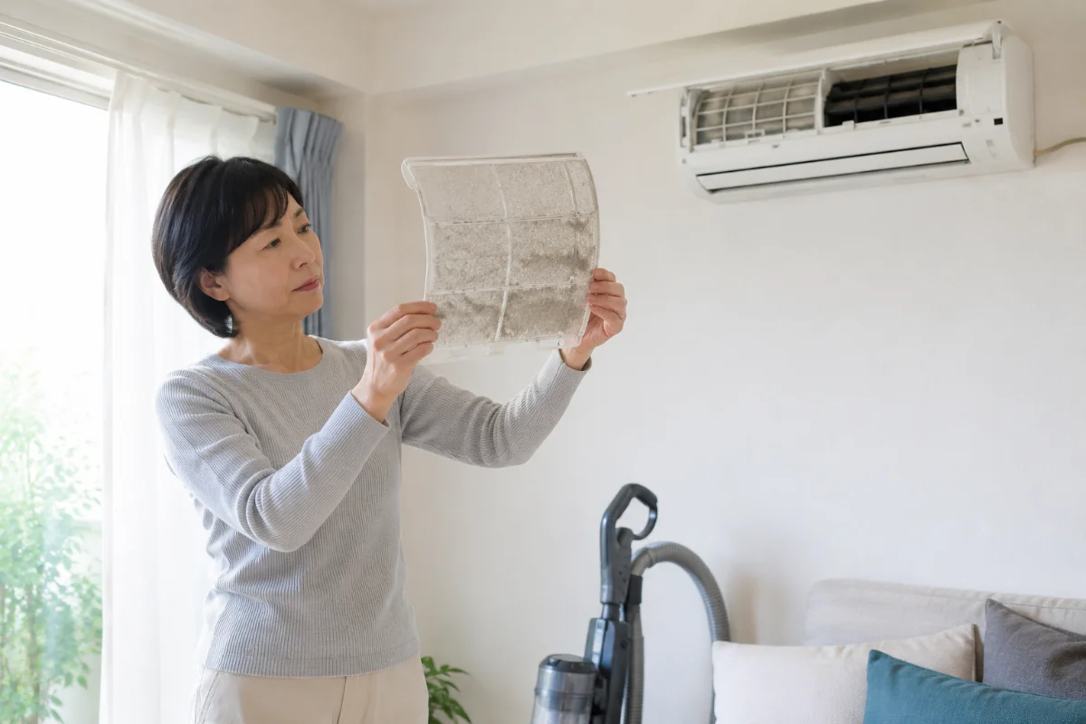 エアコンから外したフィルターのホコリの付き具合を確認する女性。そばに掃除機